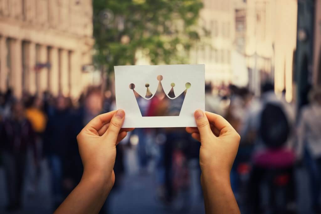 Hands holding a white paper sheet with crown icon over a crowded city street background. Business group leadership concept, power and confidence symbol.