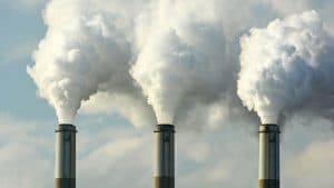 A imagem mostra três chaminés industriais emitindo grandes quantidades de fumaça branca ou vapor para o céu. As nuvens densas de emissões se espalham, contrastando com o fundo de um céu parcialmente nublado. A cena sugere poluição do ar e impactos ambientais relacionados à atividade industrial (startups, chaminés da indústria emitindo gás carbônico e poluição)