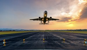 Avião decolando em uma pista de aeroporto ao pôr do sol, com o céu em tons alaranjados e montanhas ao fundo. A cena transmite movimento e a grandiosidade da aviação, aereo avião, avião decolando, decolagem, turismo, seguro