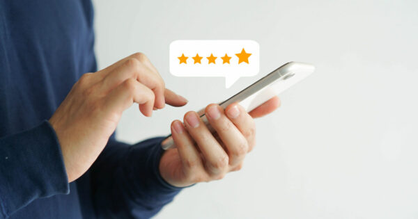 Imagem de uma pessoa segurando um smartphone com uma das mãos enquanto utiliza a outra para tocar na tela. Acima do dispositivo, há um balão de diálogo com cinco estrelas douradas, representando uma avaliação de alta qualidade ou feedback positivo. O fundo é branco, destacando a interação com o aparelho (clientes, experiência do cliente, CX)