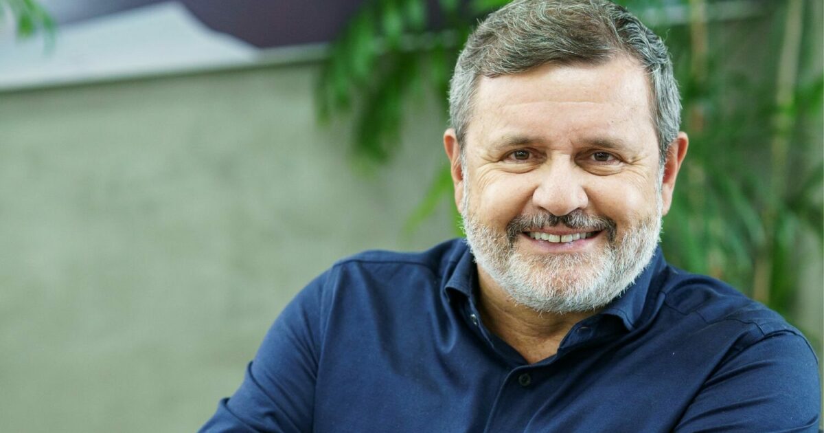 Vittorio Danesi, CEO da Simpress, sorrindo para a câmera. Ele é um homem de meia-idade com barba grisalha e cabelo curto, vestindo uma camisa azul escura. O fundo da imagem é levemente desfocado, com elementos verdes de plantas e uma parede neutra (online)