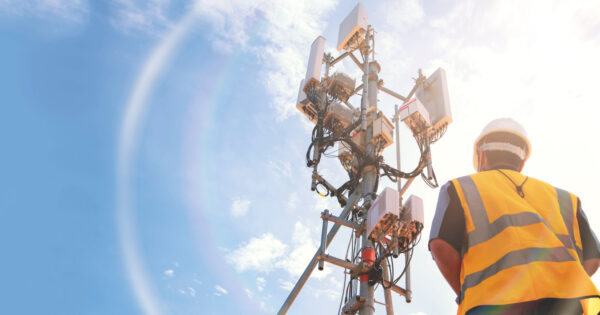 HPE Imagem de um trabalhador usando colete de alta visibilidade e capacete, olhando para uma torre de telecomunicações equipada com antenas e equipamentos de transmissão. O céu está azul com algumas nuvens, iluminado pela luz do sol. A cena representa o avanço tecnológico e a infraestrutura necessária para a conectividade 5G (redes, Anatel, 5G, ministério das comunicações, Conexis, investimentos)