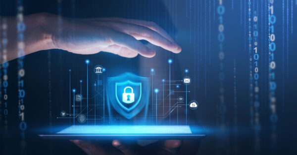 Imagem conceitual de cibersegurança, mostrando uma mão protegendo um escudo com ícone de cadeado no centro, representando proteção digital. O fundo apresenta elementos tecnológicos, como números binários e ícones de conectividade, destacando segurança em ambientes digitais (ciberataques, segurança, proteção de dados, inteligência artificial, segurança cibernética) 99