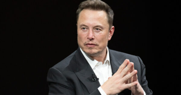 A imagem mostra Elon Musk, o CEO de empresas como Tesla, Xai, SpaceX. Ele está com cabelo curto e um leve sorriso, usando um terno escuro e uma camisa branca. Ele está sentado e posando com as mãos unidas, em um gesto de reflexão, em um fundo escuro e simples. Sua expressão facial transmite seriedade e foco, sugerindo um ambiente profissional, possivelmente durante uma entrevista ou palestra. A iluminação suave destaca seu rosto e postura (Elon Musk, dell, x, xai, starlink, spacex, tiktok)