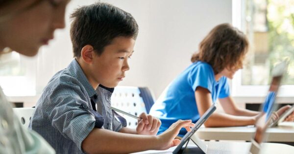 Grupo de crianças em uma sala de aula moderna, concentradas em atividades usando tablets. Em primeiro plano, um menino com expressão focada toca a tela do tablet com uma caneta digital, enquanto outras crianças ao fundo também interagem com dispositivos tecnológicos. A luz natural entra através de grandes janelas, iluminando o ambiente de aprendizado digital e colaborativo (Estudantes, escola, alunos, aprendizado, educação, tecnologia, escolas, crianças, ensino, rnp)