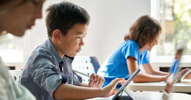 Grupo de crianças em uma sala de aula moderna, concentradas em atividades usando tablets. Em primeiro plano, um menino com expressão focada toca a tela do tablet com uma caneta digital, enquanto outras crianças ao fundo também interagem com dispositivos tecnológicos. A luz natural entra através de grandes janelas, iluminando o ambiente de aprendizado digital e colaborativo (Estudantes, escola, alunos, aprendizado, educação, tecnologia, escolas, crianças, ensino, rnp)