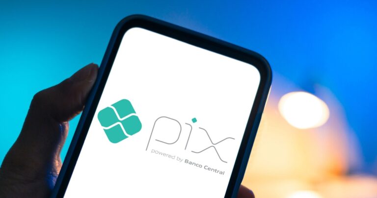 Pessoa segurando um smartphone que exibe na tela o logo do Pix, sistema de pagamento instantâneo brasileiro desenvolvido pelo Banco Central. O logotipo é composto por um símbolo geométrico verde ao lado da palavra "Pix" em letras estilizadas, seguido da frase "powered by Banco Central". O fundo da tela é branco e o ambiente ao redor tem iluminação com tons de azul e laranja desfocados (Sensedia, CNI)