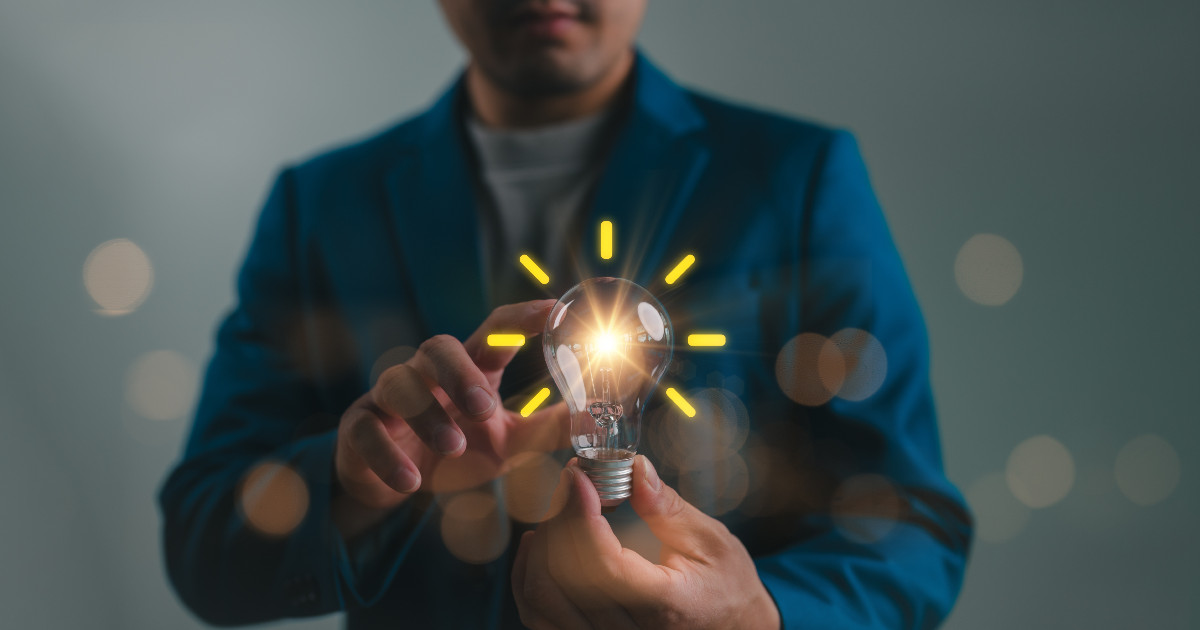 Homem segurando uma lâmpada acesa com raios de luz emanando, simbolizando inovação e criatividade. Ele veste um blazer azul em um fundo neutro, destacando o conceito de ideias brilhantes e soluções inovadoras (inovação, empresa, startup, empresas, ideia, negócios, Andrade Gutierrez, tendências, CIO)