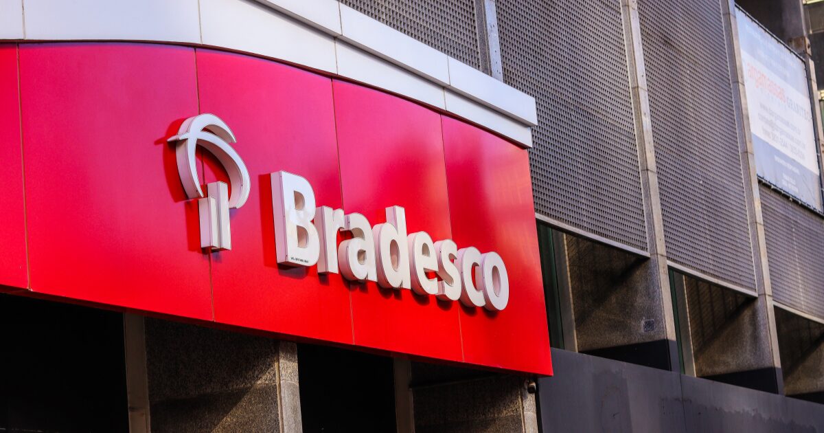 A imagem mostra a fachada de uma agência do banco Bradesco, com o logotipo característico em branco sobre um fundo vermelho. A estrutura tem um design moderno, com painéis metálicos perfurados ao fundo e um toldo curvo que abriga a identidade visual da instituição financeira (transações,voz, Fundação Bradesco)