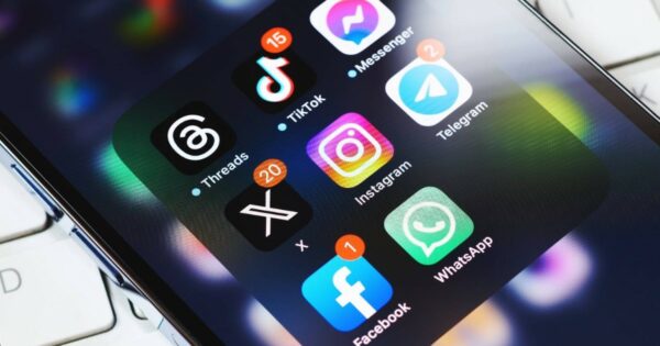 Tela de smartphone com vários aplicativos de redes sociais e mensagens abertos em uma pasta, incluindo Threads, TikTok, Messenger, Telegram, X (antigo Twitter), Instagram, Facebook e WhatsApp. Cada aplicativo exibe notificações de mensagens não lidas. A imagem está levemente inclinada, mostrando parte do teclado de um notebook ao fundo (apps, aplicativos, redes sociais, celular, smartphone, apps externos CGI, Windows), IA (ECA Digital)