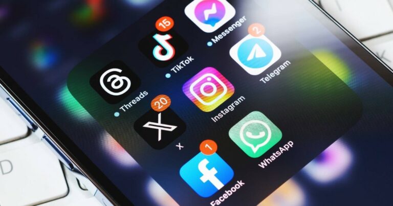 Tela de smartphone com vários aplicativos de redes sociais e mensagens abertos em uma pasta, incluindo Threads, TikTok, Messenger, Telegram, X (antigo Twitter), Instagram, Facebook e WhatsApp. Cada aplicativo exibe notificações de mensagens não lidas. A imagem está levemente inclinada, mostrando parte do teclado de um notebook ao fundo (apps, aplicativos, redes sociais, celular, smartphone, apps externos CGI, Windows), IA (ECA Digital)