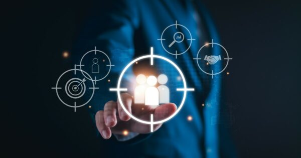 Texto alternativo: Pessoa vestindo um terno azul tocando uma tela virtual com um ícone de grupo de pessoas dentro de uma mira digital. Ao redor, há outros ícones representando alvos, análise de dados, pesquisa e aperto de mãos, simbolizando estratégias de gestão de clientes e marketing direcionado. O fundo é escuro com um efeito tecnológico e futurista. (Gestão, clientes, tecnoloiga, negócios, preparado identificar, hiperpersonalização, continuidade, Nonaka, experiência)