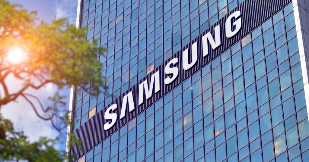 Fachada de um prédio moderno com janelas de vidro refletindo o céu, destacando o logotipo da Samsung em letras brancas. À esquerda, galhos de uma árvore com folhas verdes aparecem sob a luz do sol, criando um contraste entre a natureza e a arquitetura contemporânea (samsung, vietnã)