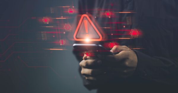 Pessoa segurando um smartphone com ícones e símbolos digitais em vermelho projetados no ar, incluindo um grande triângulo de alerta com um ponto de exclamação. A imagem simboliza um alerta de segurança cibernética ou vulnerabilidade em dispositivos móveis, com elementos gráficos digitais ao fundo (iot, ciberataque, vulnerabilidade, alerta, segurança, cibersegurança, dispositivo, device, golpe, ataques maliciosos, risco, empresa)