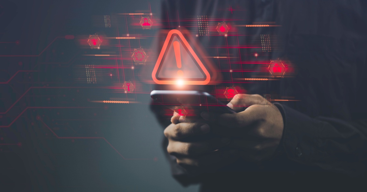 Pessoa segurando um smartphone com ícones e símbolos digitais em vermelho projetados no ar, incluindo um grande triângulo de alerta com um ponto de exclamação. A imagem simboliza um alerta de segurança cibernética ou vulnerabilidade em dispositivos móveis, com elementos gráficos digitais ao fundo (iot, ciberataque, vulnerabilidade, alerta, segurança, cibersegurança, dispositivo, device, golpe, ataques maliciosos, risco, empresa)