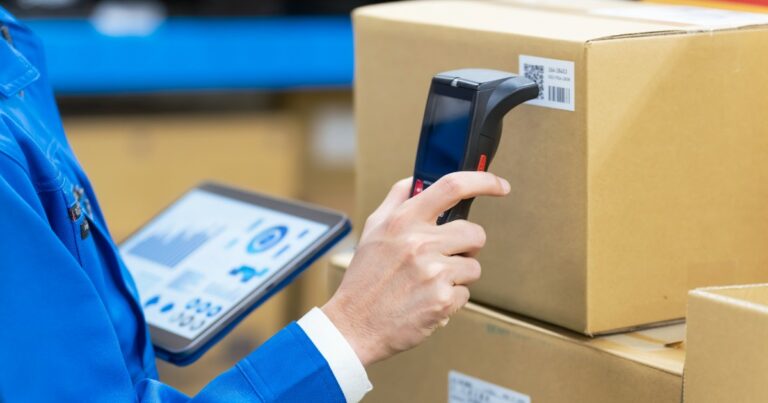 Trabalhador em um ambiente de logística utilizando um scanner de código de barras em uma caixa de papelão, enquanto segura um tablet com gráficos e dados exibidos na tela. Ele veste uniforme azul e está cercado por pacotes, sugerindo um processo de controle de estoque e rastreamento de produtos em um armazém (carbono, tecnologia)
