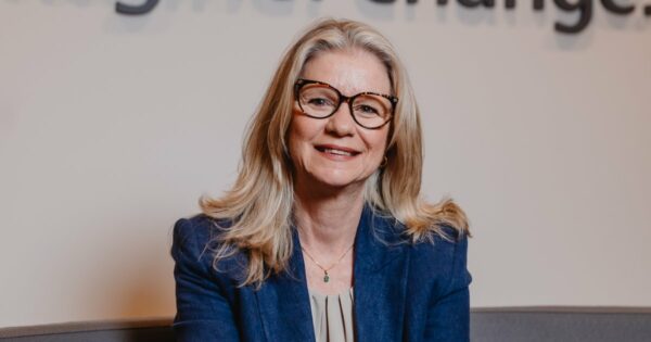 Andrea Klevenhusen, CEO da Ricoh Brasil, sorrindo enquanto está sentada. Ela usa um blazer azul e óculos de armação grande com cabelo loiro comprido e solto. O fundo é desfocado com um texto parcialmente visível