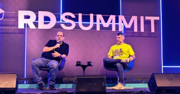 Foto de Germán Quiroga, conselheiro e investidor, sócio da Omini11 Consultoria, à esquerda, segurando um microfone enquanto fala durante o RD Summit. Ao seu lado direito, Patrick Scripilliti, especialista de digital commerce da Totvs, veste uma camiseta amarela e escuta atentamente. O letreiro iluminado 'RD Summit' e luzes de neon criam uma atmosfera vibrante no palco do evento, e-commerce