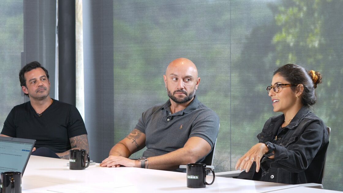 Imagem de uma mesa redonda de discussão durante a IT Forum Series, com três participantes engajados em uma conversa sobre liderança em TI. À direita, uma mulher fala enquanto os outros dois participantes, à esquerda e ao centro, a escutam atentamente. Cada participante tem uma caneca da série sobre a mesa. O fundo desfocado de tons esverdeados cria um ambiente profissional e amigável