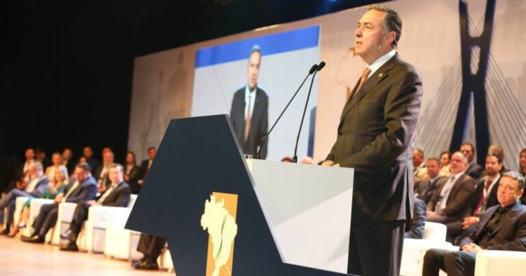 A imagem mostra um homem de terno, gravata e postura séria, discursando em um púlpito decorado com um mapa estilizado do Brasil. Ele está em um auditório, com diversas pessoas sentadas ao fundo, aparentando ser um público formal, possivelmente autoridades ou representantes de instituições. No palco, há um telão que exibe o orador, reforçando a ideia de um evento oficial ou conferência. Ao fundo, uma estrutura com design moderno, que pode ser associada à Ponte Estaiada, sugere que o evento ocorre em São Paulo ou remete à arquitetura da cidade. (STF)