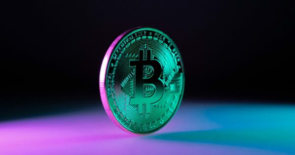 Imagem de uma moeda de Bitcoin em pé sobre uma superfície com iluminação em tons de roxo e azul. A moeda é mostrada em um tom verde metálico, destacando o símbolo do Bitcoin '₿' ao centro. O fundo está escuro, proporcionando um contraste que realça a moeda e os detalhes de sua superfície.