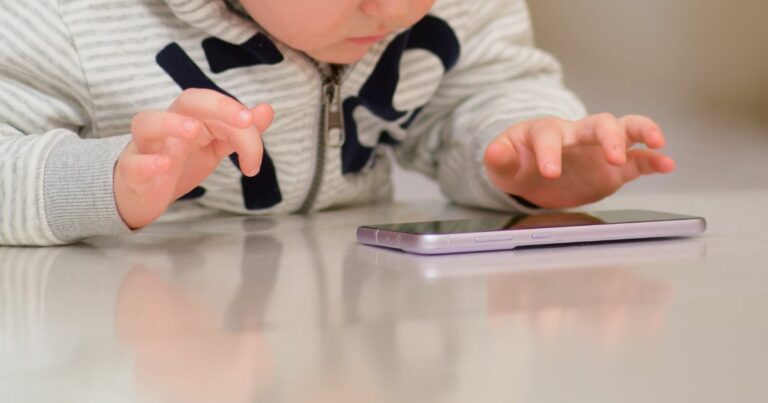 Esta imagem mostra uma criança pequena debruçada sobre uma superfície brilhante, interagindo com um smartphone. As suas mãos pequenas estão posicionadas perto do dispositivo, como se estivesse a tocar no ecrã ou a explorar o telemóvel. A criança veste um casaco cinzento com padrão listrado e um fecho de correr visível, e a cena sugere concentração ou curiosidade infantil. O foco está na interação entre a criança e o smartphone, representando um momento moderno e comum na infância atual. Kaspersky