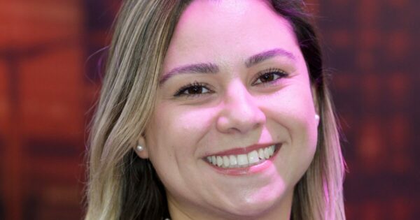 Uma mulher sorrindo com o rosto levemente iluminado por uma luz rosada. Ela tem cabelos lisos e claros, que caem sobre os ombros. Seus olhos são castanhos, e ela usa brincos de pérola. A imagem tem um fundo desfocado em tons quentes, destacando o rosto dela. Especialista da Oakmont