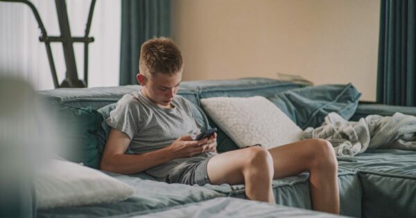 Adolescente sentado em um sofá, usando um smartphone para acessar suas redes sociais, em um ambiente doméstico com almofadas e uma manta ao fundo. (Google, Meta, Character.AI, chatgpt, segurança)