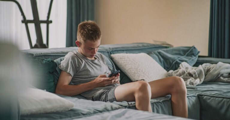 Adolescente sentado em um sofá, usando um smartphone para acessar suas redes sociais, em um ambiente doméstico com almofadas e uma manta ao fundo. (Google, Meta, Character.AI, chatgpt, segurança)