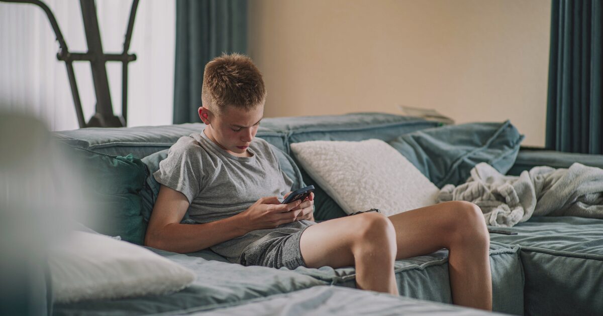 Adolescente sentado em um sofá, usando um smartphone para acessar suas redes sociais, em um ambiente doméstico com almofadas e uma manta ao fundo. (Google, Meta, Character.AI, chatgpt, segurança)