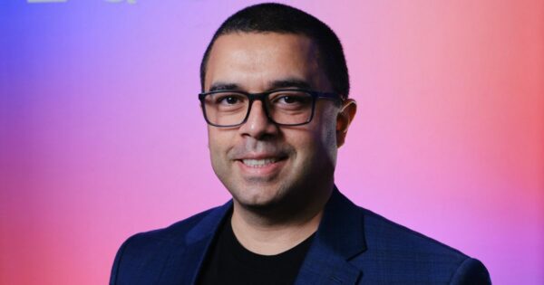 Victor Arnaud, executivo da Equinix, sorrindo para a câmera com um fundo de cores vibrantes em tons de roxo e rosa. Ele usa óculos e veste um blazer azul escuro sobre uma camiseta preta, transmitindo um visual moderno e profissional