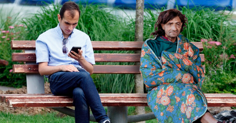 Homem sentado em um banco de parque, olhando para o celular, enquanto ao seu lado uma pessoa sem-teto está sentada envolta em um cobertor floral, destacando o contraste entre realidades sociais trabalho