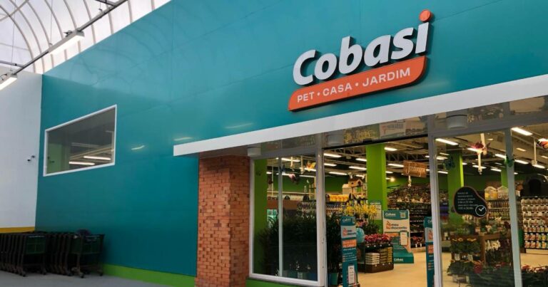 A imagem mostra a fachada de uma loja da Cobasi, uma rede de varejo especializada em produtos para pets, casa e jardim. A entrada é iluminada, com uma grande janela de vidro que revela o interior da loja, que parece ser organizada e bem decorada. O nome "Cobasi" está em destaque, em letras grandes e brancas, acompanhado dos termos "PET · CASA · JARDIM", com a palavra "Cobasi" destacada em laranja e verde. Em frente à loja, há alguns carrinhos de compras empilhados. A decoração interna parece ter várias prateleiras com produtos para pets e jardinagem