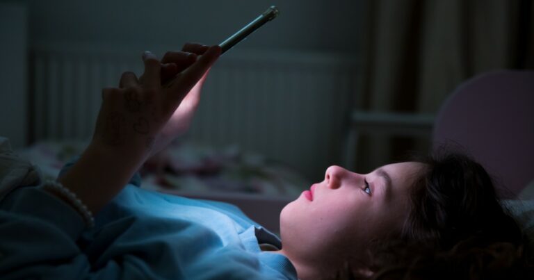 A imagem mostra uma criança deitada na cama, usando um smartphone à noite. A luz da tela do celular ilumina suavemente seu rosto, enquanto ela observa o dispositivo com atenção. A criança está usando uma blusa de cor azul e possui tatuagens temporárias visíveis em uma das mãos. O ambiente ao fundo é sombrio, com pouca iluminação, destacando a luz emitida pelo celular. A cena transmite a ideia de que a criança está interagindo com a tecnologia, possivelmente em uma rede social ou aplicativo, Australia