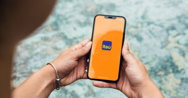Pessoa segurando um smartphone com a tela exibindo o logotipo do Itaú sobre um fundo laranja. A imagem destaca o uso do aplicativo bancário, simbolizando praticidade e tecnologia, com um fundo desfocado que dá foco à interação com o dispositivo (itau, itau unibanco, app, superapp, inteligencia itau, código)