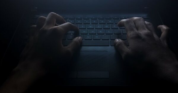 Mãos digitando em um teclado de laptop em um ambiente escuro, iluminadas por uma luz fraca que destaca o cenário misterioso e sombrio. A imagem evoca o conceito do lado obscuro da tecnologia, como cibercrimes ou atividades clandestinas, valor, tsmc