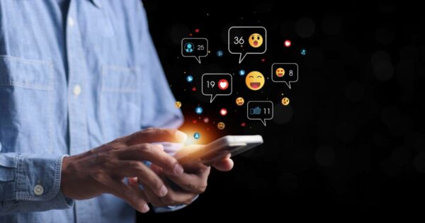 Pessoa utilizando um smartphone enquanto ícones de interações digitais, como curtidas, comentários e reações com emojis, flutuam ao redor da tela. O fundo escuro destaca a luminosidade dos ícones, representando o engajamento em redes sociais e o impacto da tecnologia na comunicação, abx, hiperpersonalização ( IA generativa), ABM, (BRQ), profissionais