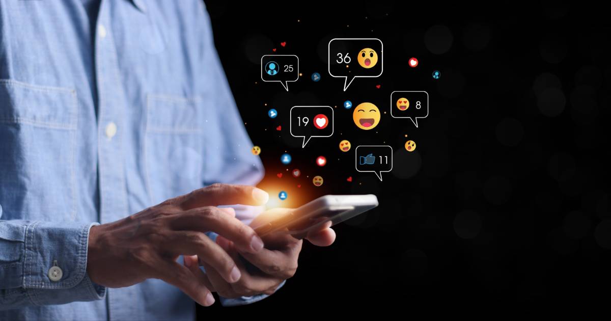Pessoa utilizando um smartphone enquanto ícones de interações digitais, como curtidas, comentários e reações com emojis, flutuam ao redor da tela. O fundo escuro destaca a luminosidade dos ícones, representando o engajamento em redes sociais e o impacto da tecnologia na comunicação, abx, hiperpersonalização ( IA generativa), ABM, (BRQ), profissionais