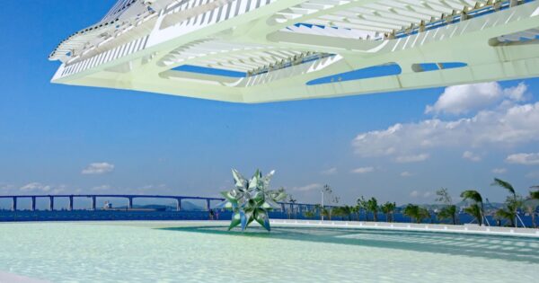 Vista externa do Museu do Amanhã, no Rio de Janeiro, com sua arquitetura futurista e estrutura branca sobre uma piscina espelhada. Ao fundo, é possível ver o céu azul e a Baía de Guanabara, incluindo a Ponte Rio-Niterói. No centro da imagem, há uma escultura geométrica refletindo luz, criando um ambiente moderno e tecnológico