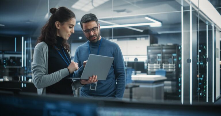 Dois profissionais de TI discutindo enquanto analisam informações em um laptop dentro de um data center moderno. O ambiente é iluminado por luzes azuis e cercado por servidores de alta tecnologia, com um cenário futurista e profissional, executivos, salários, empregos (Archimate, desenvolvedor)