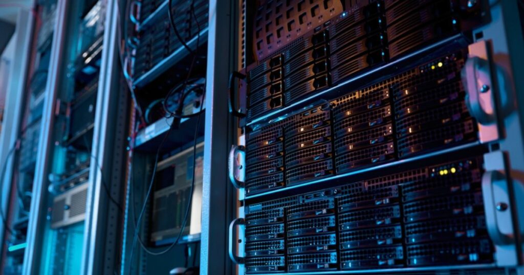 A imagem mostra um ambiente de data center com diversos servidores montados em racks. Os equipamentos estão organizados em fileiras verticais, com luzes indicadoras acesas em alguns dos dispositivos, sugerindo que estão em operação. A iluminação é predominantemente azulada, com tons sutis de roxo, criando uma atmosfera tecnológica e moderna. Cabos e componentes conectam os servidores, refletindo a infraestrutura robusta necessária para armazenamento e processamento de dados em larga escala. Esse cenário é típico de locais utilizados para operações de TI críticas, como hospedagem de sites, análise de dados, inteligência artificial e outras aplicações de computação avançada. (emissões, brasil, tecto, CLM)