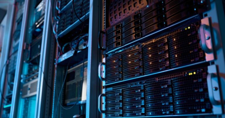 A imagem mostra um ambiente de data center com diversos servidores montados em racks. Os equipamentos estão organizados em fileiras verticais, com luzes indicadoras acesas em alguns dos dispositivos, sugerindo que estão em operação. A iluminação é predominantemente azulada, com tons sutis de roxo, criando uma atmosfera tecnológica e moderna. Cabos e componentes conectam os servidores, refletindo a infraestrutura robusta necessária para armazenamento e processamento de dados em larga escala. Esse cenário é típico de locais utilizados para operações de TI críticas, como hospedagem de sites, análise de dados, inteligência artificial e outras aplicações de computação avançada. (emissões, brasil, tecto)