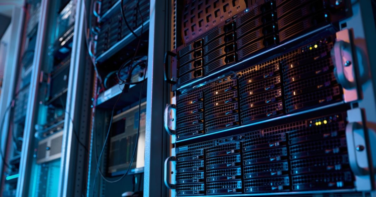 A imagem mostra um ambiente de data center com diversos servidores montados em racks. Os equipamentos estão organizados em fileiras verticais, com luzes indicadoras acesas em alguns dos dispositivos, sugerindo que estão em operação. A iluminação é predominantemente azulada, com tons sutis de roxo, criando uma atmosfera tecnológica e moderna. Cabos e componentes conectam os servidores, refletindo a infraestrutura robusta necessária para armazenamento e processamento de dados em larga escala. Esse cenário é típico de locais utilizados para operações de TI críticas, como hospedagem de sites, análise de dados, inteligência artificial e outras aplicações de computação avançada. (emissões, brasil, tecto)