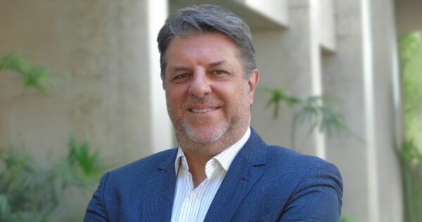 Dennis Milan, diretor de tecnologia, inovação e digital da Tokio Marine, sorrindo em um ambiente externo com plantas e paredes claras ao fundo. Ele está vestido com um blazer azul e camisa branca, transmitindo profissionalismo e confiança
