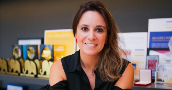 Uma mulher sorridente está sentada, olhando diretamente para a câmera. Ela tem cabelos castanhos claros, parcialmente soltos, e usa brincos vermelhos chamativos. Está vestida com uma blusa preta sem mangas. Ao fundo, há várias prateleiras decoradas com troféus dourados e placas de premiação, sugerindo um ambiente profissional e de conquistas.