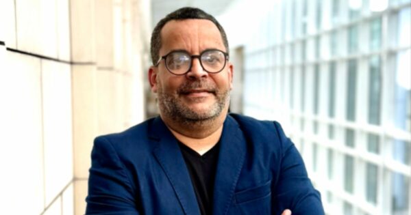 Um homem de pele clara, usando óculos de armação redonda, veste um blazer azul e está posicionado em frente a uma estrutura moderna de vidro e concreto. Ele sorri levemente, transmitindo confiança. A imagem ilustra um contexto sobre agentes de IA e inovação tecnológica.