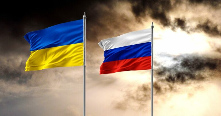 Bandeiras da Ucrânia e da Rússia hasteadas lado a lado contra um céu nublado, simbolizando o contraste entre os dois países no cenário atual de tensões geopolíticas e conflitos. A imagem reflete um momento de incertezas e desafios globais na guerra, que agora usa robôs armados