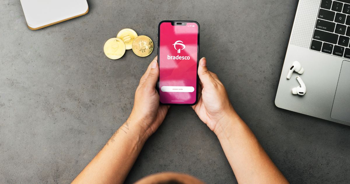 Uma pessoa segura um smartphone com o aplicativo Bradesco aberto na tela, exibindo o logotipo e um campo para login. O telefone está sobre uma mesa cinza, ao lado de três moedas físicas representando criptomoedas, incluindo Bitcoin. No canto direito, há um laptop prateado parcialmente visível com um par de AirPods ao lado. O ambiente é minimalista e sugere um contexto de finanças ou tecnologia, pix, plataforma