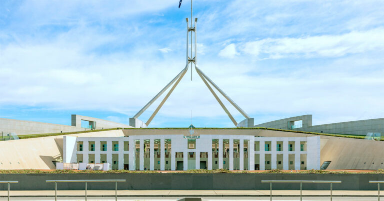 Fachada do Parlamento Australiano em Canberra, destacando sua arquitetura moderna com linhas simétricas, colunas brancas e um grande mastro central com a bandeira da Austrália. O céu azul com nuvens complementa a vista frontal do edifício, redes sociais