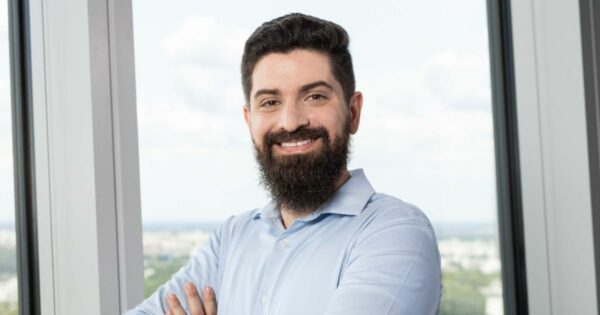 Thiago Bertacchini, head de vendas da Mangopay, sorrindo com os braços cruzados em um ambiente corporativo com janelas amplas ao fundo. Ele veste uma camisa azul clara, transmitindo confiança e profissionalismo